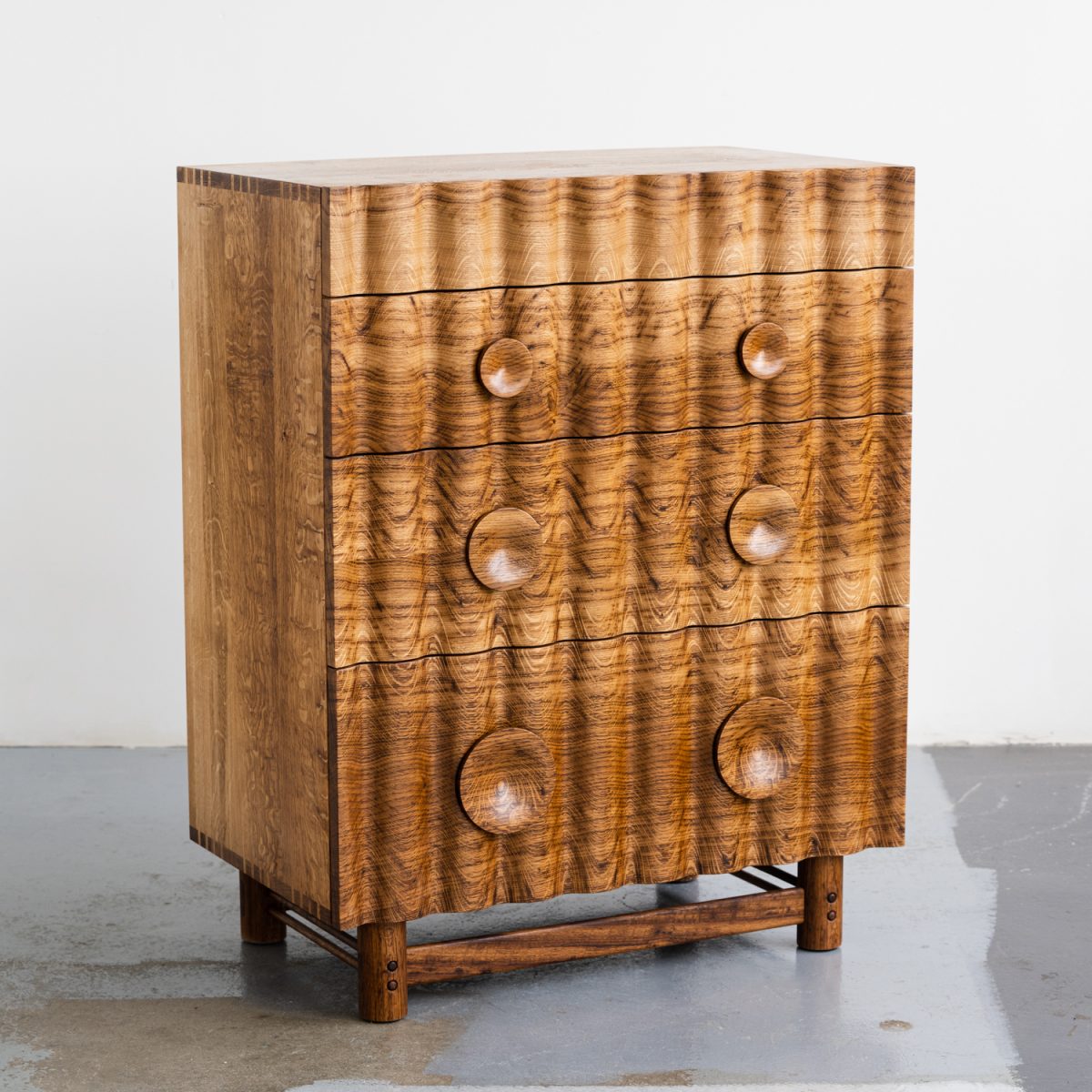 Bowater Chest of Drawers, a tiered four drawer unit. The signature ripple facade is digitally profiled from brown oak, hand-turned leg and handle details with a removable base, The carcass is made from solid timber with dovetail joinery