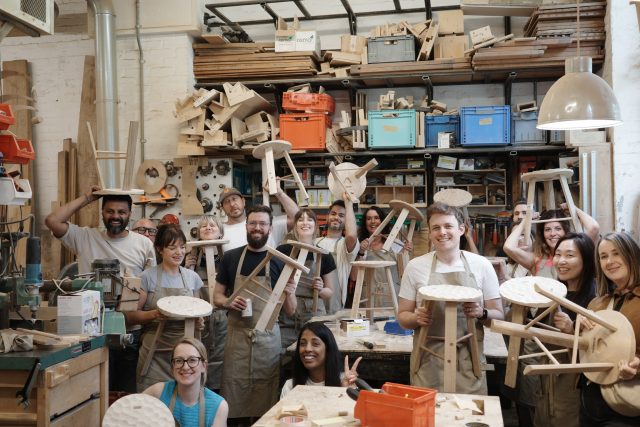 Large group of 24 workshop participants smiling and cheering while holding wooden stools above their heads in a craft workshop.