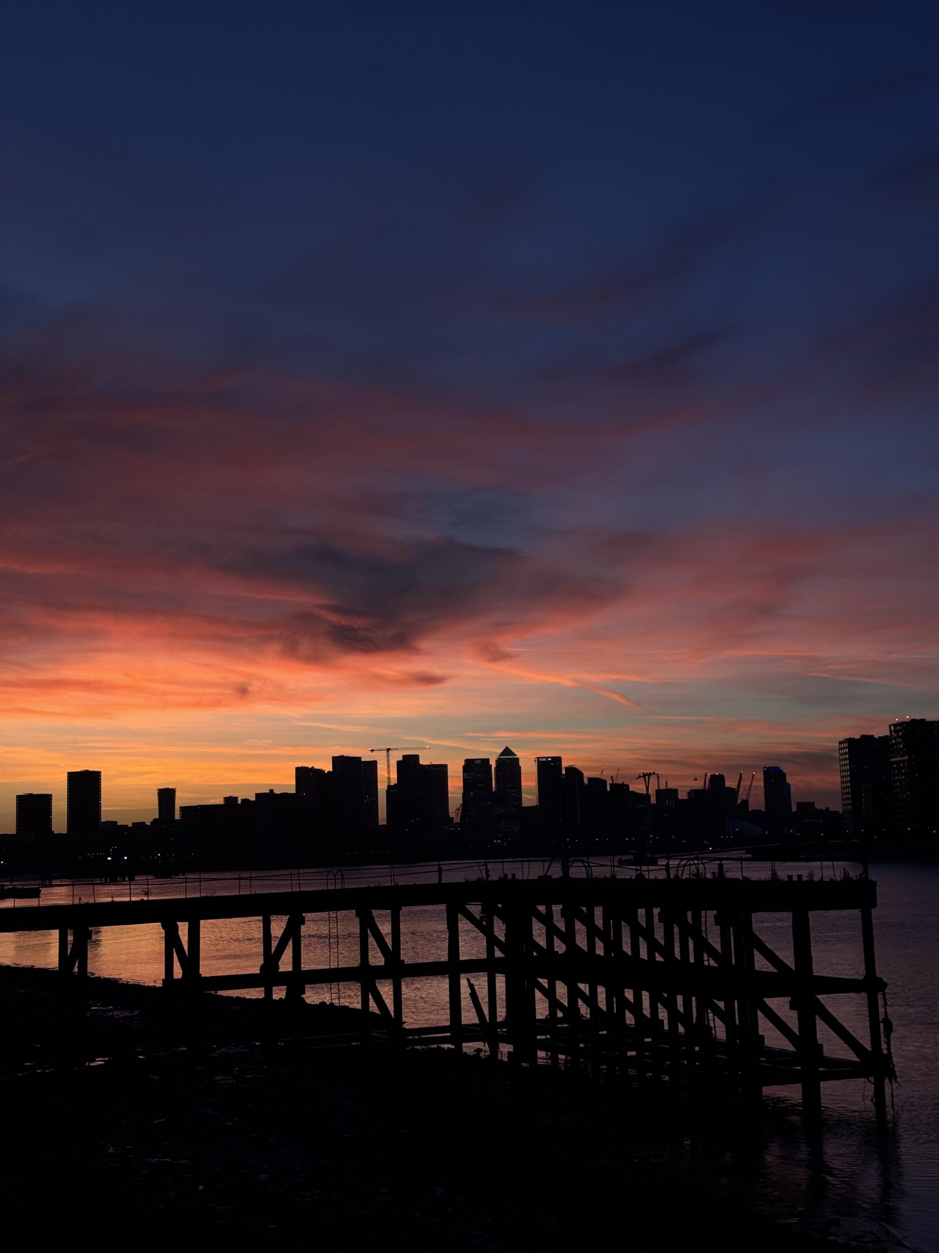 Early autumn sunset over the River Thames from Woolwich, with an old timber jetty in silhouette and the Canary Wharf skyline beyond.